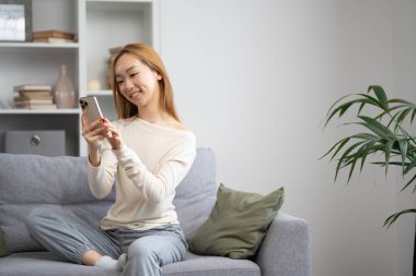 Mutlu Asyalı Kadın Gri Koltukta Akıllı Telefon Kullanıyor, Günlük Yaşam Tarzı, Modern Oturma Odası, İçerik Yaratıcısı, Evdeki Teknoloji