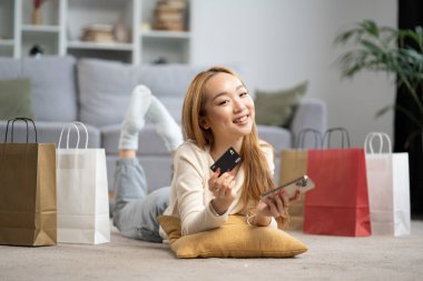Evde alışveriş yapan mutlu genç bir kadın kredi kartı ve cep telefonu ile alışveriş torbaları tutuyor. E-ticaret, çevrimiçi alışveriş, tüketim kavramı.
