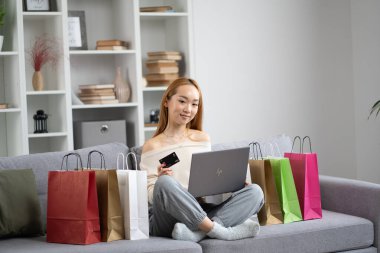Çevresinde renkli alışveriş torbaları, modern yaşam tarzı ve e-ticaret konsepti olan renkli mobilyalarla çevrimiçi alışveriş yapan mutlu genç kadın.