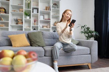 Genç bir kadın evdeki koltukta eğleniyor, rahat, rahatlık ve modern yaşam tarzıyla rahat oturma odasında akıllı telefona gülümsüyor.