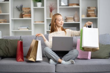Genç Kadın, çevrimiçi alışveriş başarısını dizüstü bilgisayarında, rahat evinde renkli alışveriş torbalarıyla çevreledi. E-ticaret kavramı, Mutlu, Memnuniyet.