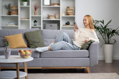 Genç bir kadın akıllı telefonuyla koltukta dinleniyor, rahat oturma odasında sosyal medyanın tadını çıkarıyor. Milenyum Yaşam Tarzı, Konforlu Alan, Modern İç Tasarım.