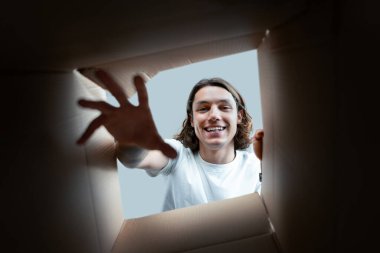 Joyful Man Unpacking Box, Smiling Young Adult Gently Reaching Out From Inside A Cardboard Box, Emphasizing Surprise And New Beginnings In A Home Setting clipart