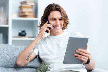 Genç adam tablet tutarken telefonda konuşmaktan keyif alıyor, modern bir oturma odasında oturuyor, mutluluk ve günlük yaşam tarzını yayıyor. Telekomünikasyon ve Teknoloji temaları için mükemmel.