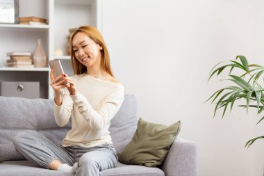 Mutlu Asyalı Kadın Gri Koltukta Akıllı Telefon Kullanıyor, Günlük Yaşam Tarzı, Modern Oturma Odası, İçerik Yaratıcısı, Evdeki Teknoloji