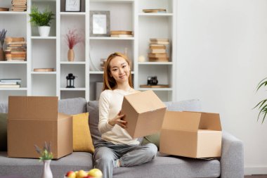 Yeni apartmanda kutuları açan genç bir kadın, kameraya mutlu bir şekilde gülümseyen, modern oturma odası arka planı. Ev, Taşınma Günü, Yeni Başlangıçlar.