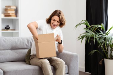 Heyecanlı genç adam evdeki koltukta gizemli paketi açıyor, neşe ve beklenti sergiliyor, e-ticaret, teslimat ve sürpriz konsept için mükemmel..