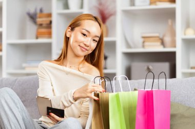 Alışveriş çantaları ve kredi kartı ile mutlu genç bir kadın, evde alışveriş yapıyor, rahat oturma odası ayarlıyor, e-ticaret anlayışı var.