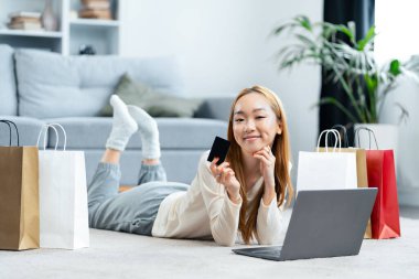 Çevrimiçi alışveriş yapmaktan hoşlanan genç bir kadın dizüstü bilgisayarla ve kredi kartıyla yalan söyleyen, sıradan bir ev hayatı yaşayan. Mutlu, Boş, E-Ticaret Konsepti.