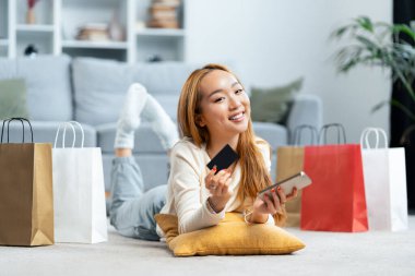 Evde alışveriş yapan mutlu genç bir kadın kredi kartı ve cep telefonu ile alışveriş torbaları tutuyor. E-ticaret, çevrimiçi alışveriş, tüketim kavramı.