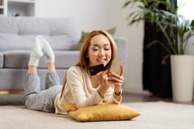 Evde akıllı telefondan alışveriş yapan, gülümseyen Asyalı kadın kredi kartı tutan, rahat bir şekilde yalan söyleyen, modern oturma odası düzenleyen genç bir kadın..