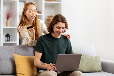 İnternetten alışveriş yapan mutlu çift. Bir erkek dizüstü bilgisayar kullanır ve bir kadın kredi kartı tutar, rahat bir oturma odasında heyecan ve neşe ifade eder..