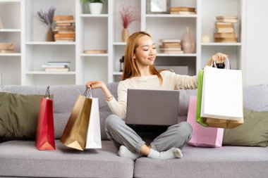 Genç Kadın, çevrimiçi alışveriş başarısını dizüstü bilgisayarında, rahat evinde renkli alışveriş torbalarıyla çevreledi. E-ticaret kavramı, Mutlu, Memnuniyet.
