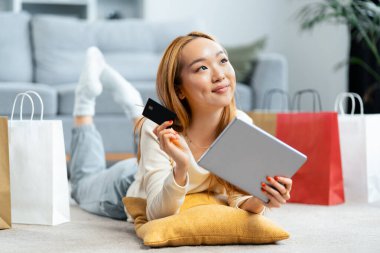 Çevrede alışveriş yapan mutlu genç kadın, kredi kartı ve dijital tablet tutuyor, alışveriş torbaları ve konforlu e-ticaret deneyimi var.