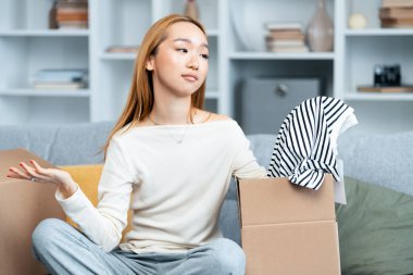 Young Woman Unpacking Clothes, Expressing Disappointment Over Fashion Purchase In Modern Living Room, Returns And Exchanges Concept clipart