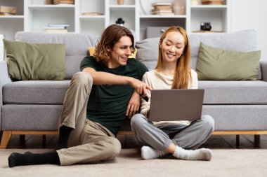 Mutlu çift evde eğleniyor, yerde oturan genç adam ve kadın rahat oturma odasında dizüstü bilgisayar kullanıyor, rahat bir hayat sürüyor.