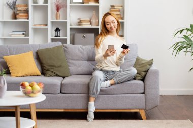Akıllı telefon ve kredi kartıyla koltukta rahatça giyinen, modern yaşam tarzını ve online alışveriş tarzını sergileyen genç bir kadın..