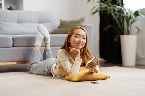 Young Woman Relaxing Home Floor Smiling While Using Smartphone Cozy Stok Foto Bebas Royalti