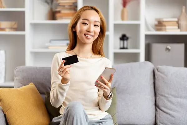 Young Asian Woman Credit Card Smartphone Sitting Sofa Smiling Camera lizenzfreie Stockbilder