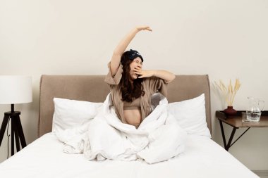 Genç bir kadın yatakta esneyerek ve esneyerek uyanıyor, rahat yatak odası ortamında, ev hayatı tarzında yenilenmiş bir sabah rutini yaşıyor.