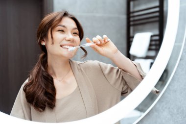 Banyodaki aynanın önünde diş fırçalayan mutlu Asyalı kadın, kişisel bakım ve hijyen yansımaları, neşeli bir sabah rutinini aydınlatan doğal ışık...