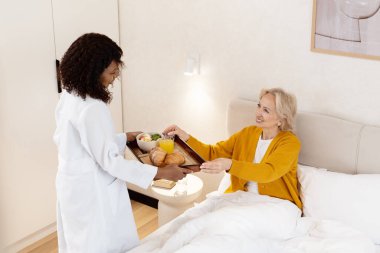 Hemşire, yatakta gülümseyen yaşlı kadına sağlıklı kahvaltı tepsisi sunuyor. Bakım, konfor ve sağlık hizmetleri sunuyor.
