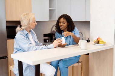 Hemşire, mutfakta kıdemli kadınla ilaçları tartışıyor, sağlık hizmetlerini, desteği ve güveni vurguluyor.