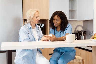 Genç bir hemşire mutfağında yaşlı bir kadına yardım ediyor, ilgi ve diyalog anlarını paylaşıyor, sağlık hizmetlerinde şefkat ve desteği ön plana çıkarıyor..