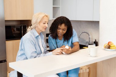 Hemşire ve kıdemli kadın evde ilaçlarını tartışıyor, bakım, destek ve modern mutfak düzenine güveniyorlar.