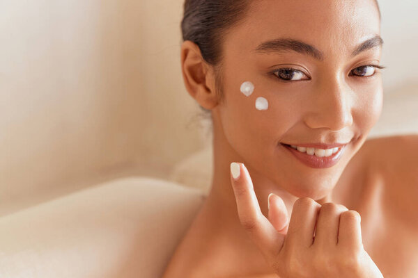 Красавица по коже. Женщина наносит крем для лица и улыбается в помещении, программа по уходу за кожей