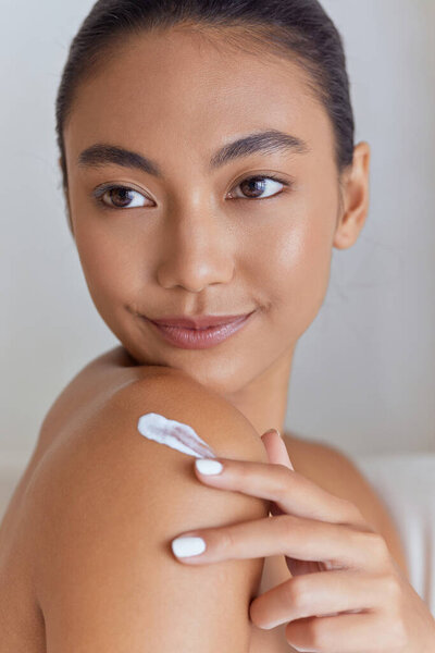 Молодая женщина наносит увлажняющий крем на плечо, содействуя здоровой коже с естественной красотой