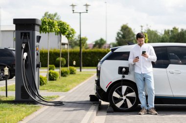 Elektirikli arabayı şarj eden ve akıllı telefon kullanan bir adam.