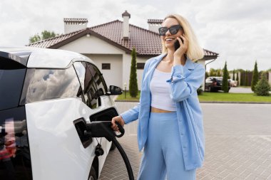 Kadın telefonda konuşurken elektrikli arabayı şarj ediyor.
