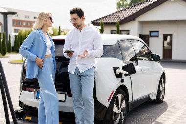 Şarj istasyonunda elektrikli arabanın yanında Eco-Friendly Taşımacılık ve Modern Yaşam tarzını vurgulayan bir çift. Parlak, güneşli bir gün, resmin olumlu ve sürdürülebilir atmosferini arttırır.
