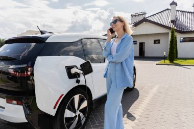 Mavi elbiseli kadın telefonla konuşurken elektrikli araba şarj ediyor, modern yaşam tarzını yansıtıyor ve çevre dostu bir yaşam sürüyor..