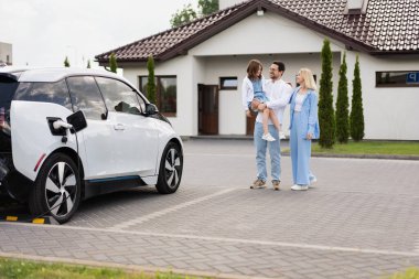 Modern bir evin önünde elektrikli arabalarının yanında duran bir aile. Kadın gülümseyip, aile bağlarını ve yeşil enerjiyi vurgularken, adam bir çocuk tutuyor.,