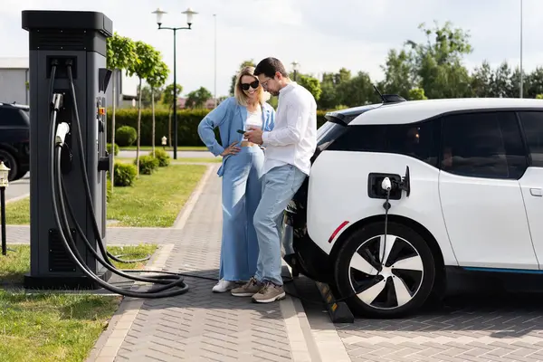 stock image Young Couple Charging Electric Car At Charging Station, Checking Mobile Phone, Embracing Sustainability And Eco-Friendly Lifestyle, Outdoors On Sunny Day