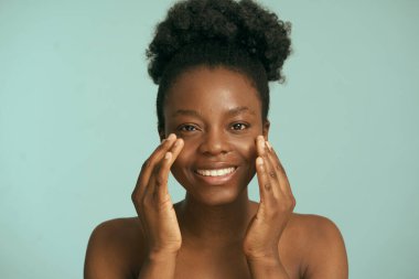 Cilt Ürünü Uygulayan Kadın. Işıl ışıl bir gülüşü ve sağlıklı parlak bir cildi olan güzel bir genç kadın. Elleri nazikçe yüzüne dokunuyor, cilt bakımını vurguluyor.. 