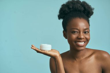 Mavi arka plana karşı cilt kremi tutan, güzelliği ve kendine özgüveni destekleyen bir kadın.