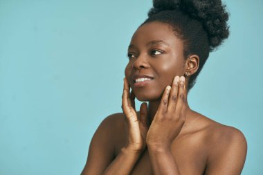 Afro Kadın iki eliyle yüzüne dokunuyor, kendine güveni ve doğal güzelliği mavi bir arka plana yansıtıyor.