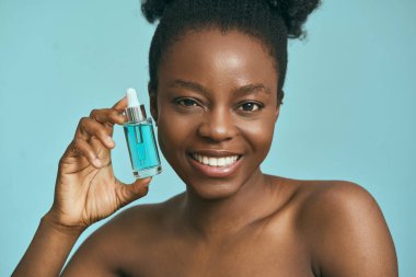 Mavi Serum Şişesine Karşı Mavi Arkaplan, Cilt Bakımı, Güzellik ve Kendine Bakım 'ı destekleyen Kadın. Özgüven, Doğal Güzellik ve Sağlıklı Cilt Duyguları vurgulanır.