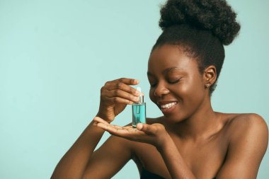 Açık mavi bir arkaplan karşısında gülümseyen, cilt bakımı ve cilt bakımı kavramlarını açığa çıkaran kadın damlatıcıyla yüz hidratımı uyguluyor