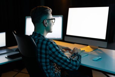 Karanlık ofiste bilgisayarda geç saatlere kadar çalışan, odaklanmış, üretken, gözlüklü ve damalı tişörtlü genç adam.