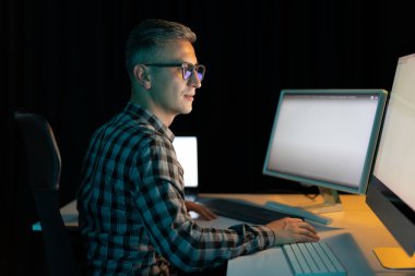 Gözlüklü adam geç saatlere kadar çalışıyor, birden fazla bilgisayar monitörüyle, konsantre ve görevine odaklanmış.