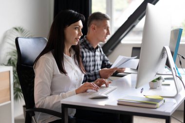 Modern bir ofis ortamında bilgisayarlar üzerinde çalışan, belgelere ve dijital görevlere odaklanan iki profesyonel. Ortak Çalışma Alanı, Takım Çalışması Kavramı
