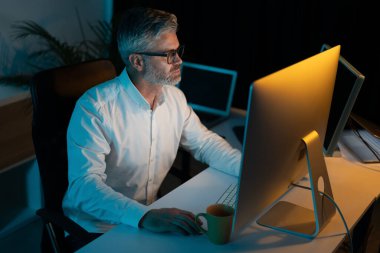 Modern ofiste bilgisayara geç saatlere kadar çalışan, çalışkan profesyonel, gece vardiyası, teknoloji ve adanmışlığa odaklanmış adam.