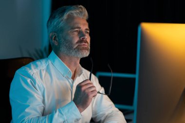 Odaklanmış son sınıf öğrencisi gece geç saatlerde bilgisayar ekranı izliyor, beyaz gömlek giyiyor ve gözlük takıyor, sıkı çalışma kavramı, derin düşünce ve konsantrasyon kavramı var.