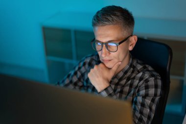 Bilgisayarda geç saatlere kadar çalışan, gözlüklü ve ekose gömlekli, modern ofis ortamında profesyonelliği ve bağlılığı yansıtan, sakin ve konsantre bir atmosfer yaratan orta yaşlı bir adam.