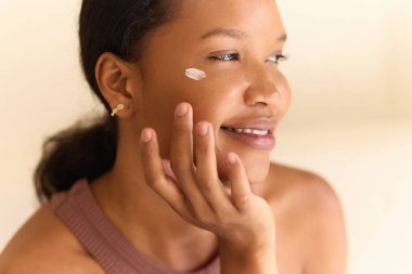 Yüz kremi uygulaması: Gülümseyen bir kadın yanağına nazikçe nemlendirici krem sürüyor, cilt bakımı, kendine bakım ve gevşemeyi vurguluyor.