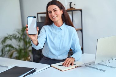 Smartphone with Blank Screen 'i gösterirken gülümseyen profesyonel iş kadını, dizüstü bilgisayarlı ve defterli ofis masasında oturuyor. Teknoloji, İletişim ve Profesyonelliği Destekliyor 